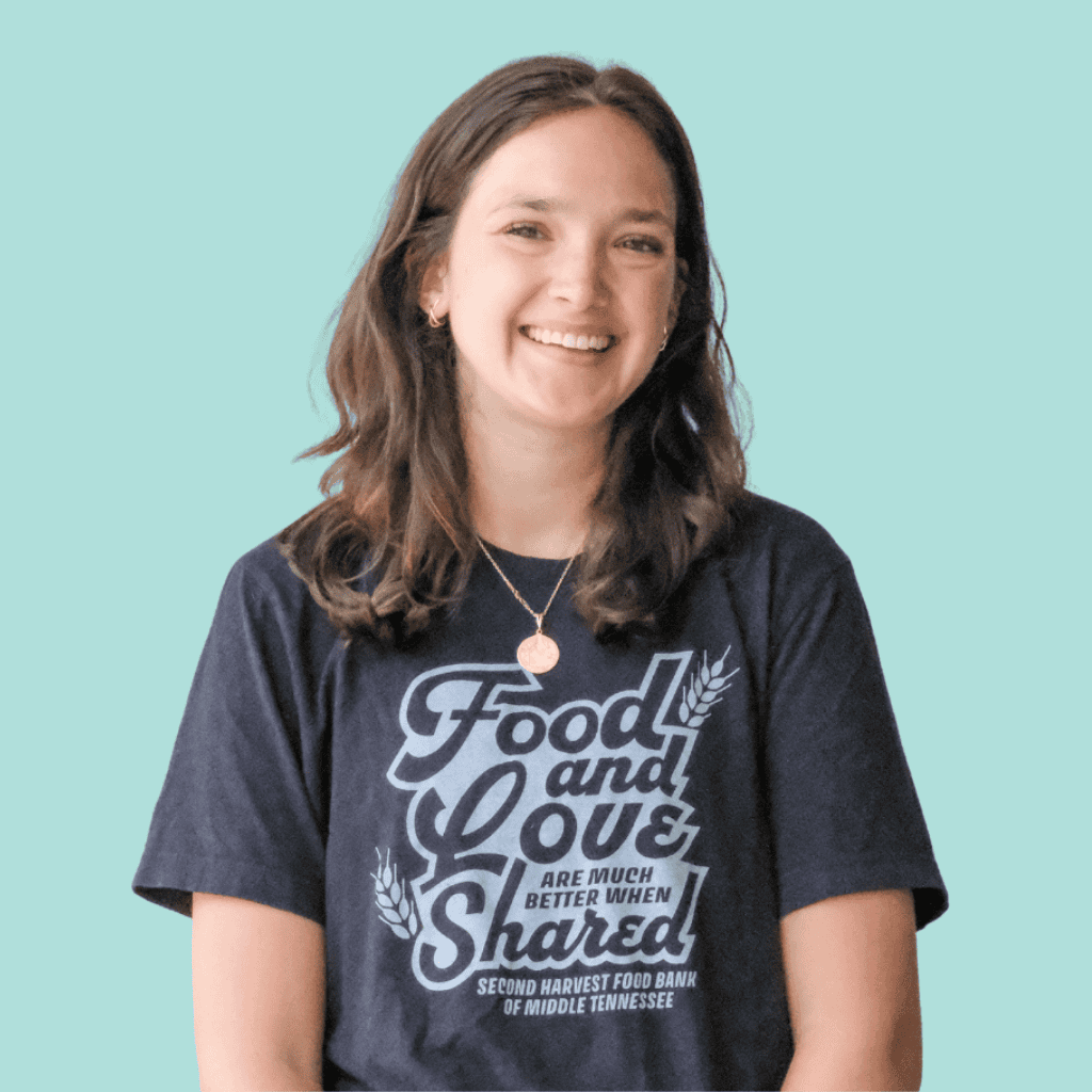 A young woman with long brown hair smiles while wearing a navy blue T-shirt that reads, “Food and Love are much better when shared—Second Harvest Food Bank of Middle Tennessee.” She stands against a light blue background.