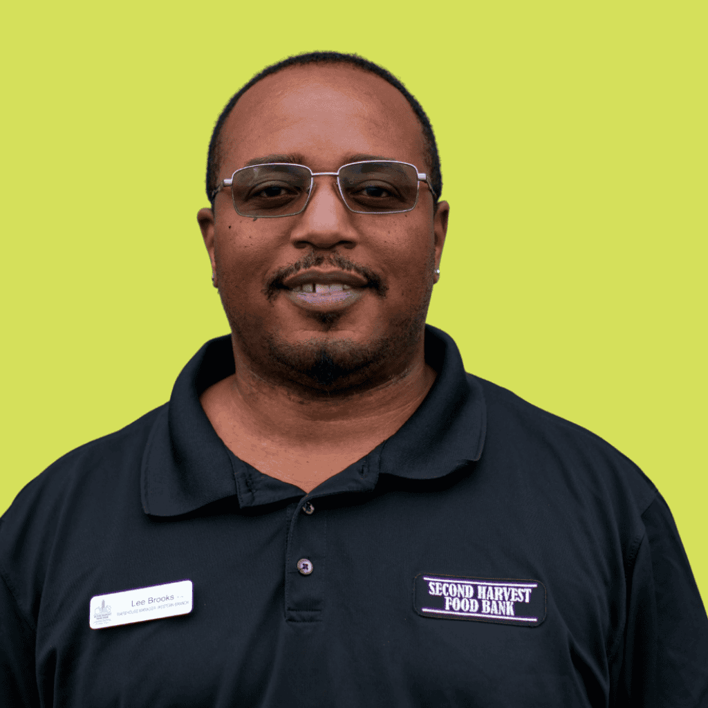 A man wearing sunglasses and a black Second Harvest Food Bank polo shirt stands against a solid light green background. He has short hair, a goatee, and a name badge that reads Lee Brooks.