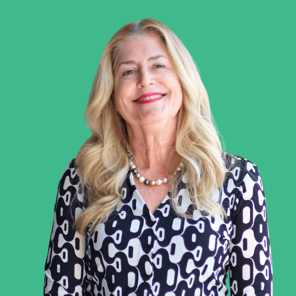 Smiling woman with long blonde hair wearing a patterned black and white blouse and a pearl necklace, standing in front of a solid teal background.