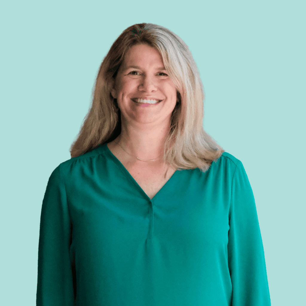 A woman with long blonde hair, wearing a teal blouse, stands smiling against a light blue background.