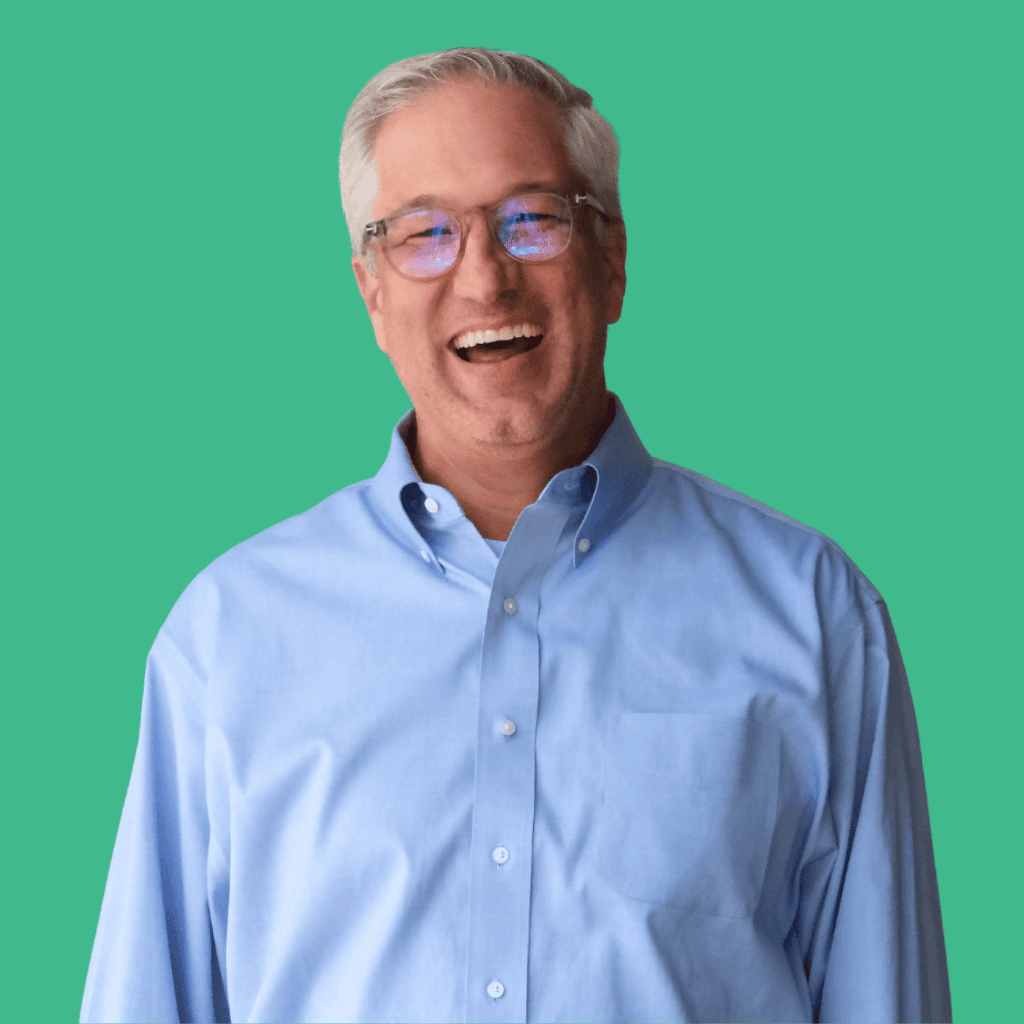 A smiling man with gray hair and glasses wearing a light blue button-up shirt stands against a solid teal background.