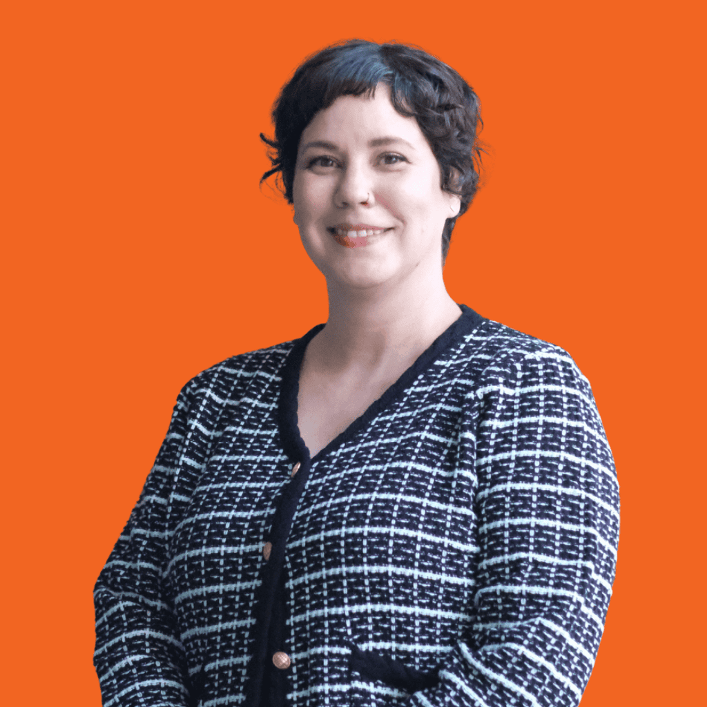 A person with short, dark curly hair is smiling and wearing a black and white checkered cardigan. The background is solid orange.