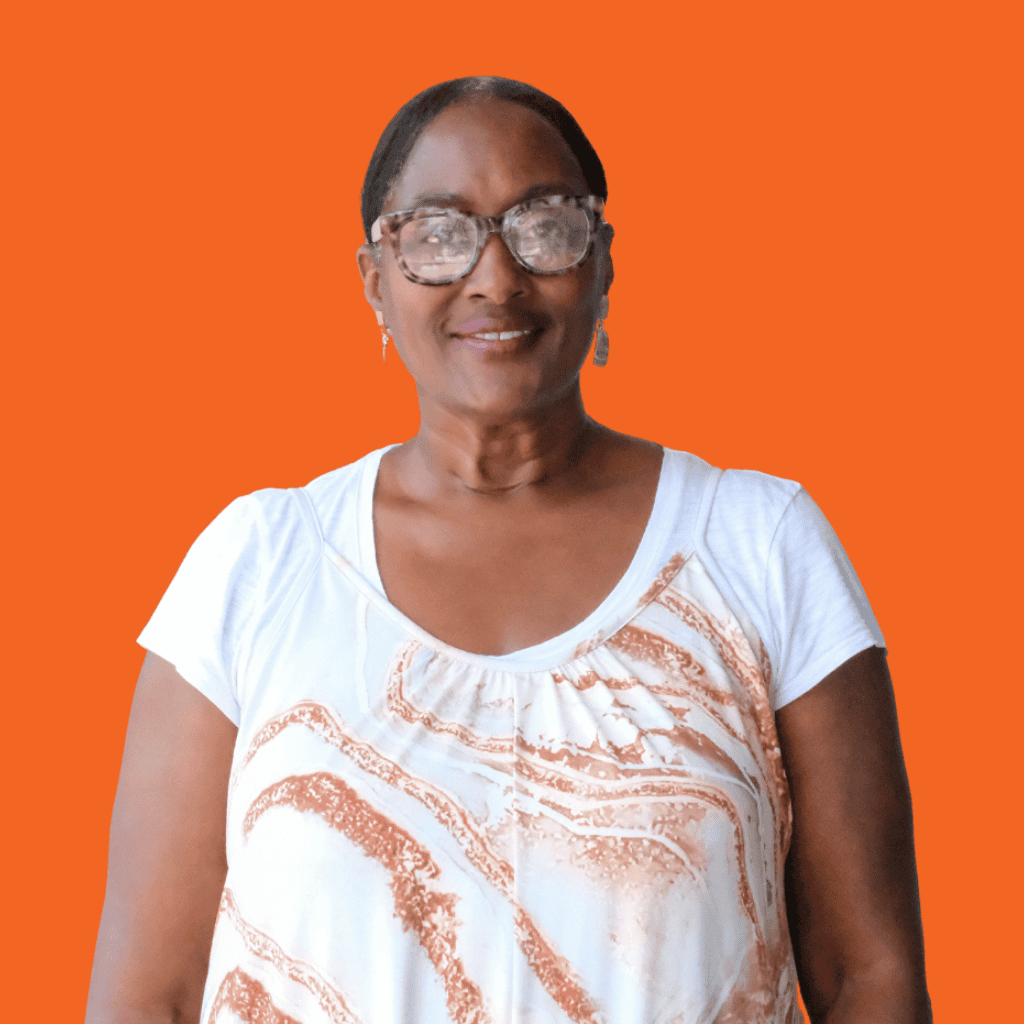A woman with glasses and earrings, wearing a white top with an orange pattern, stands smiling in front of a solid orange background.