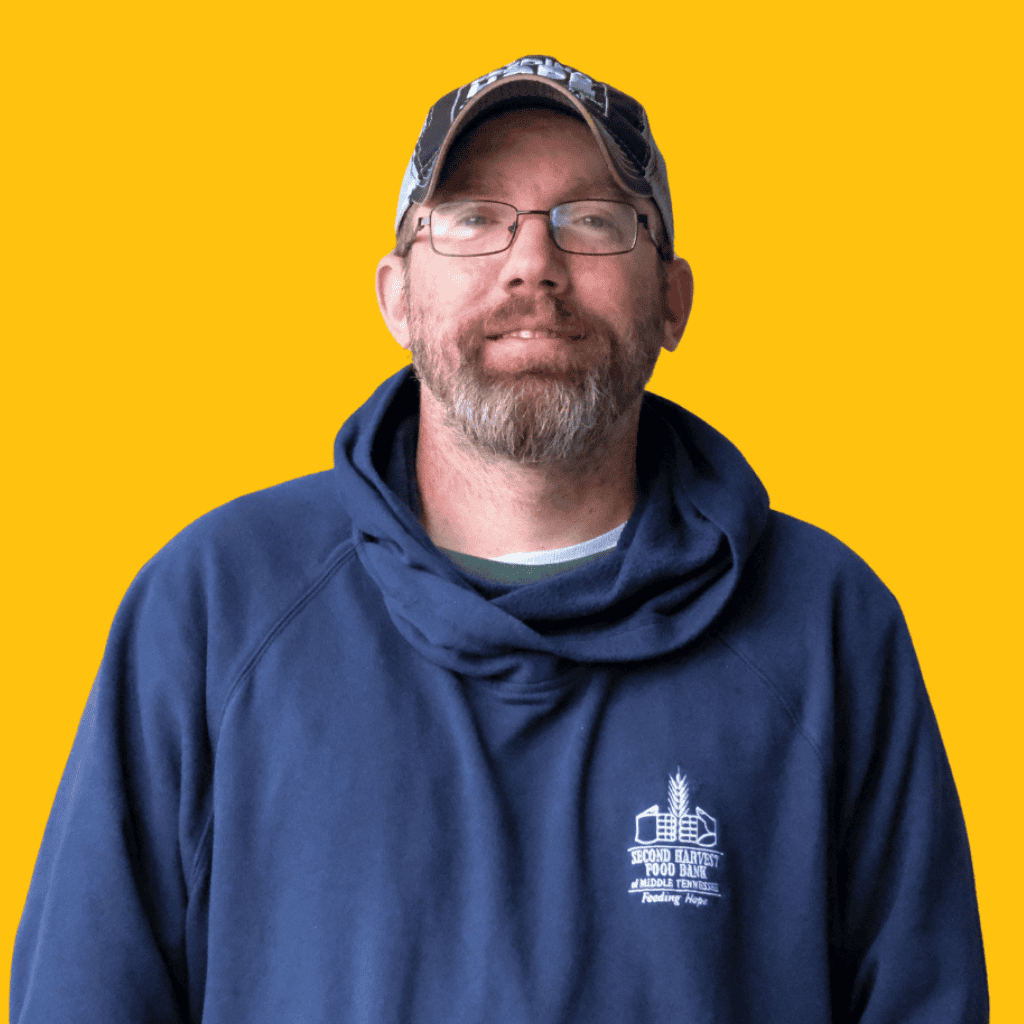 A man with a beard and glasses wearing a blue hoodie and a baseball cap stands against a solid yellow background.
