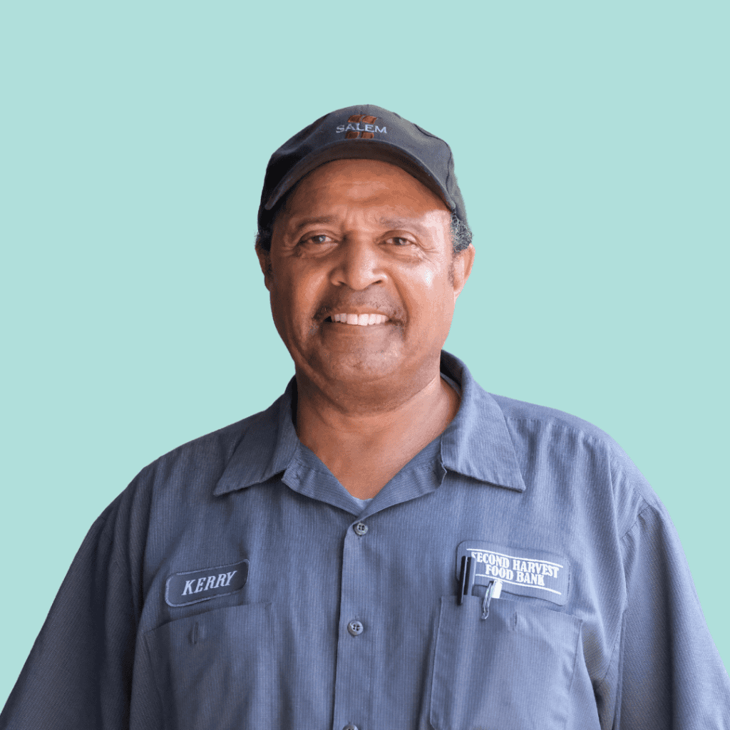 A smiling man wearing a dark baseball cap and a blue work shirt with a Second Harvest Food Bank logo and a name tag that says Kerry. He stands in front of a plain light blue background.