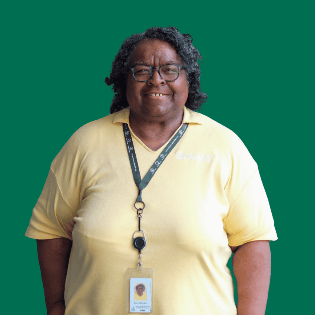 A person with curly hair and glasses, wearing a yellow shirt and a lanyard with an ID badge, stands smiling against a solid green background.