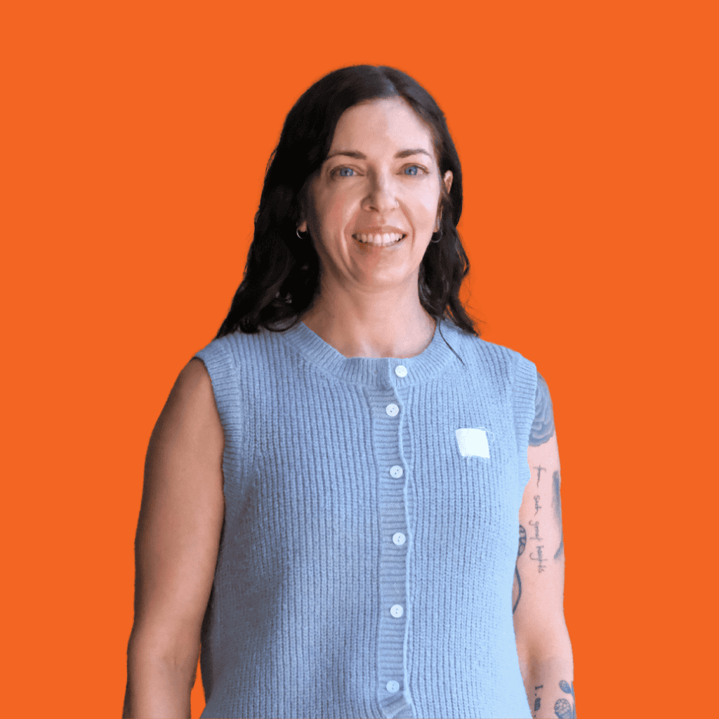 A woman with long dark hair, wearing a sleeveless light blue buttoned sweater, smiles against a solid orange background. She has tattoos on her left arm.