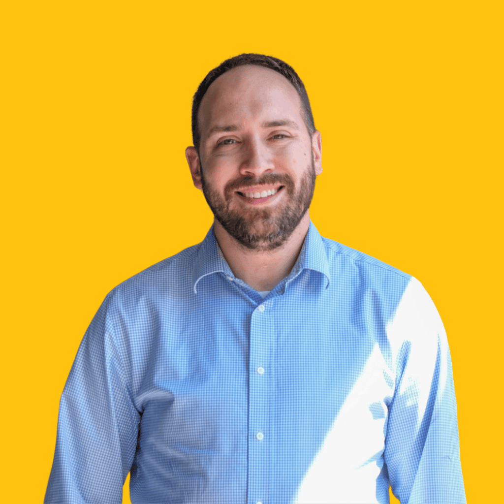 A man with short brown hair and a beard, wearing a light blue button-up shirt, smiles while standing against a bright yellow background.