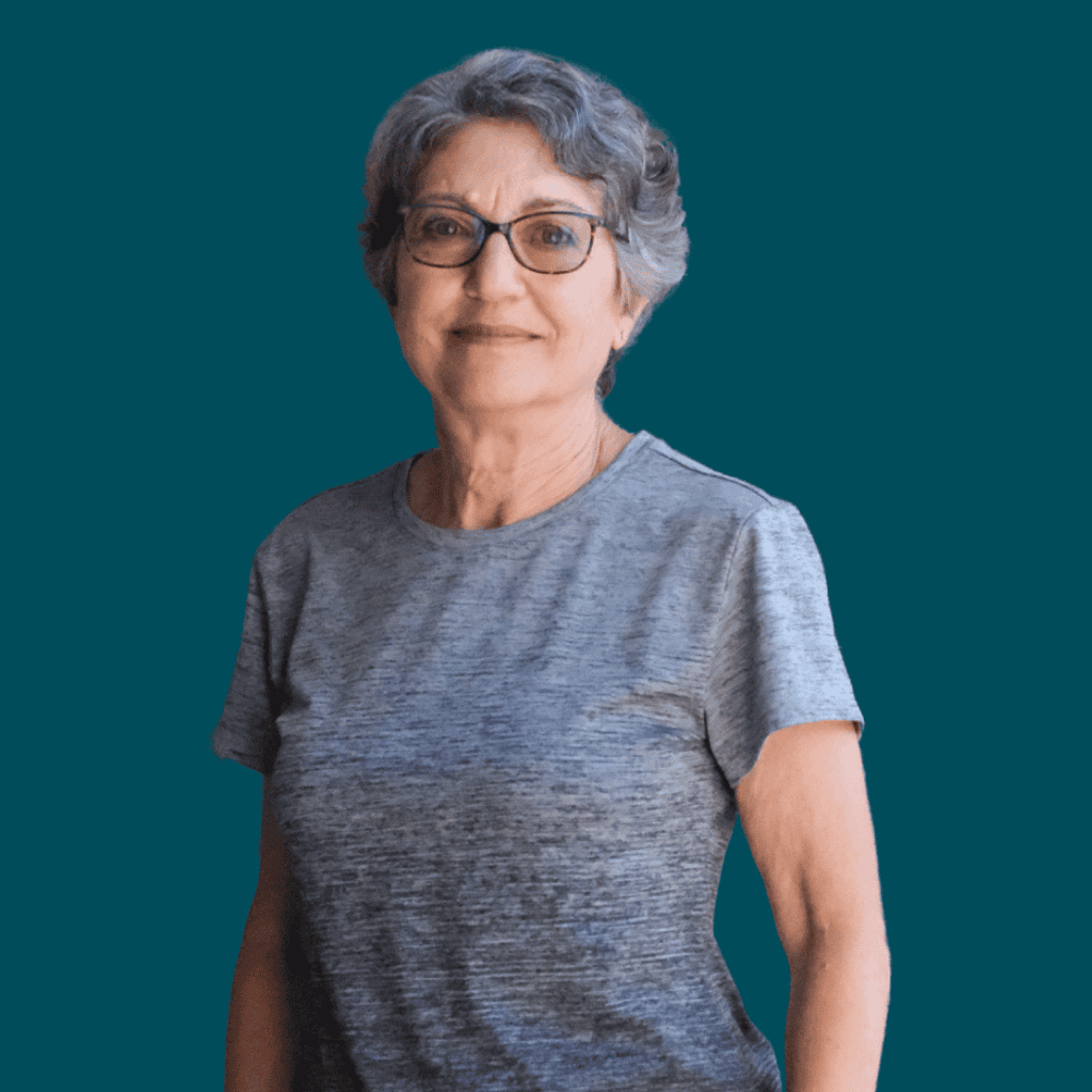An older woman with short gray hair and glasses is smiling, wearing a gray short-sleeve shirt, standing against a solid teal background.