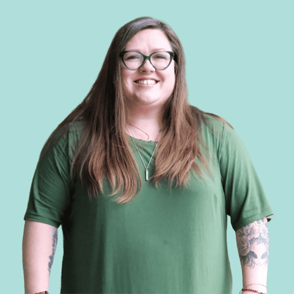 A smiling woman with long brown hair and glasses, wearing a green shirt and a necklace, stands in front of a light teal background. She has tattoos on both forearms.