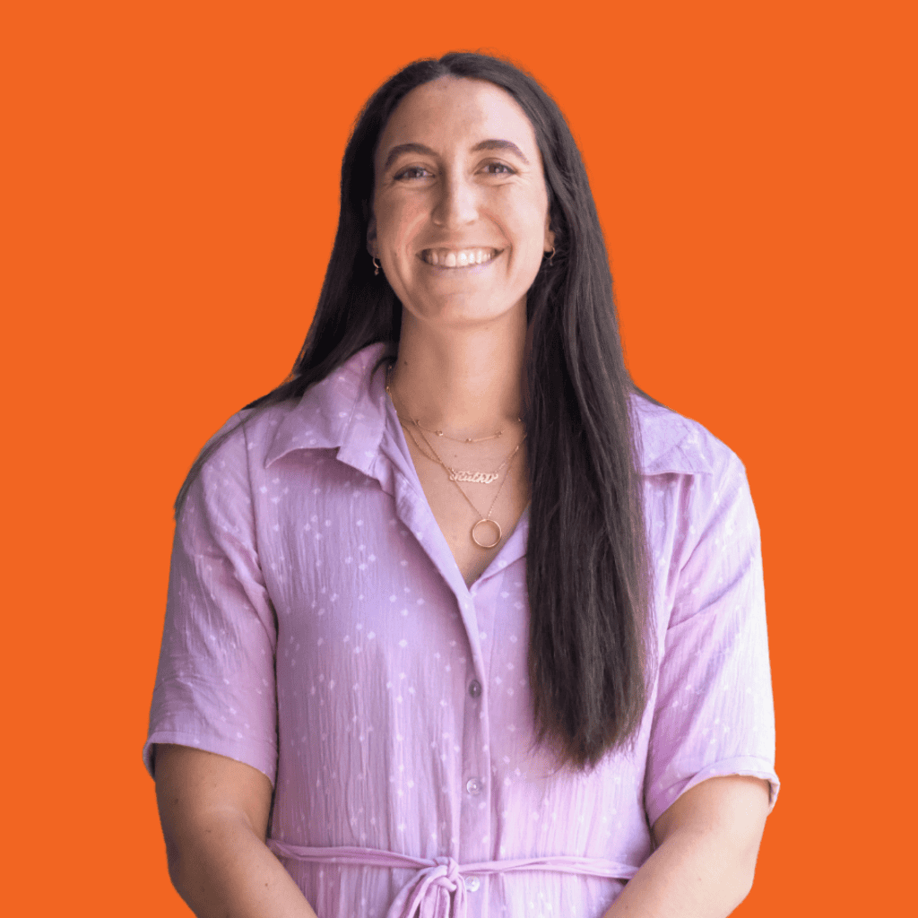 A woman with long dark hair, wearing a light purple dress and layered necklaces, smiles while standing in front of a solid orange background.