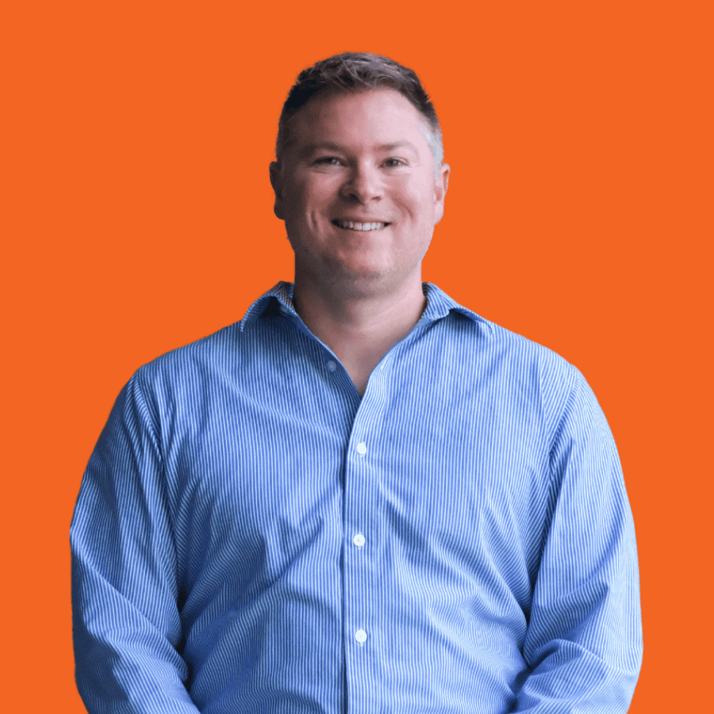 A man wearing a blue and white striped button-up shirt is smiling while standing against an orange background.