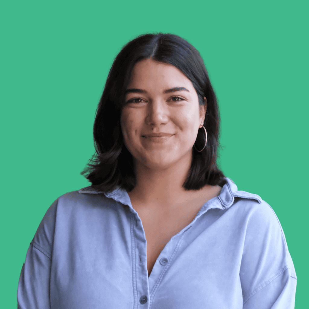 A woman with shoulder-length dark hair, wearing hoop earrings and a light blue button-up shirt, smiles softly while standing against a solid green background.