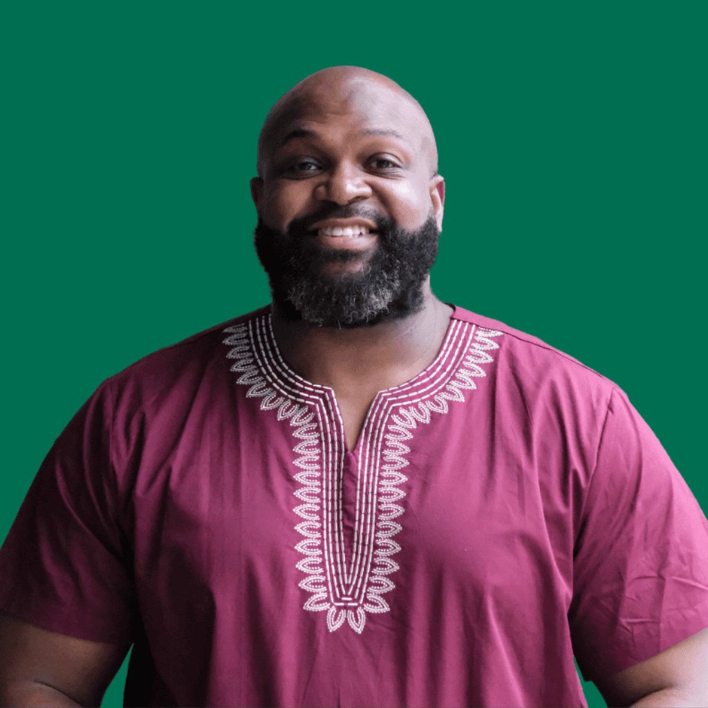 A smiling man with a beard wears a maroon shirt with white embroidered patterns, standing in front of a plain green background.