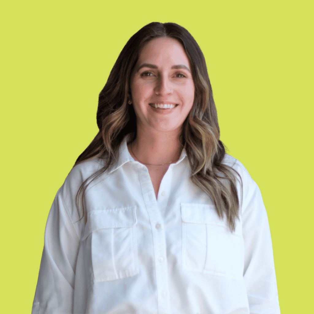 A woman with long brown hair wearing a white button-up shirt smiles at the camera. She is standing against a solid light green background.