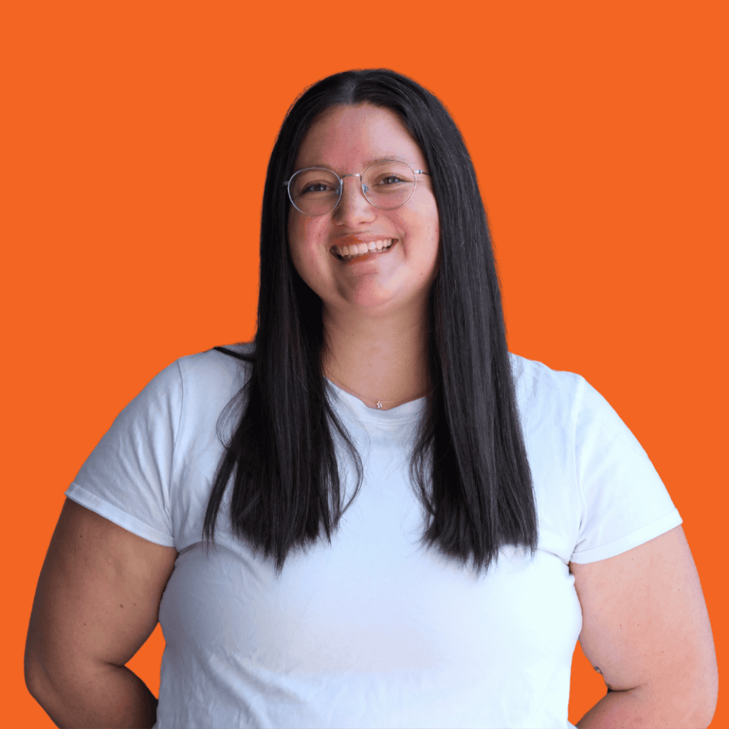 A woman with long straight dark hair, wearing glasses and a white t-shirt, is smiling while standing against an orange background.