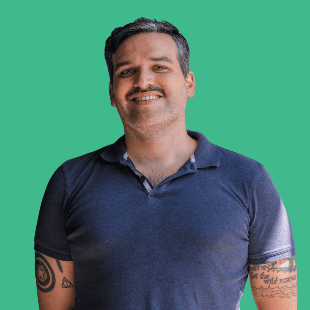 A smiling man with short dark hair and a mustache, wearing a dark blue polo shirt, stands against a solid green background. He has visible tattoos on both arms.
