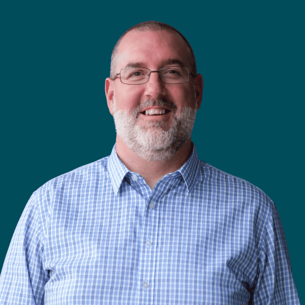 Smiling man with glasses, a short beard, and a balding head, wearing a blue and white checked button-down shirt, standing against a solid teal background.