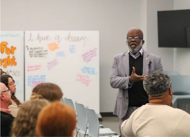 A man in a gray suit gestures while speaking to a seated audience in a classroom. Behind him, a whiteboard displays handwritten notes and quotes. People listen attentively.
