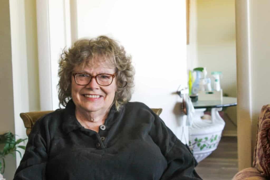 An older woman with curly gray hair and glasses smiles while sitting indoors, wearing a dark shirt. There is a plant and some cleaning supplies visible in the background.