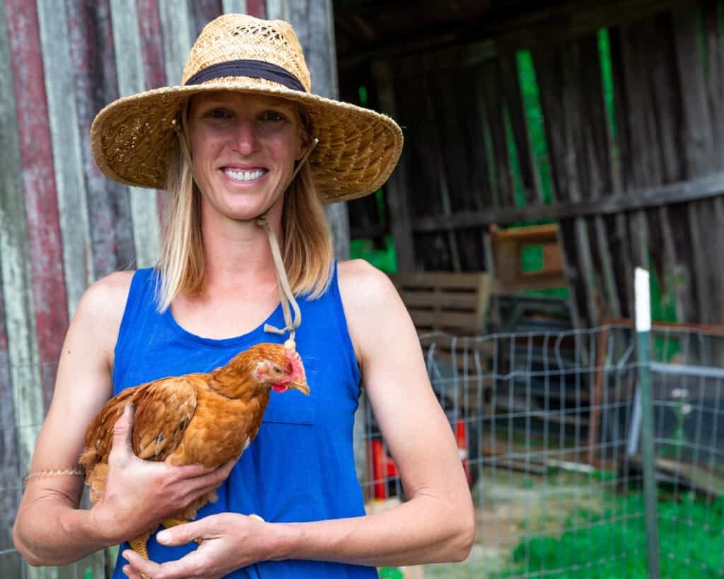 A smiling woman in a straw hat and blue tank top holds a brown chicken while standing in front of a rustic wooden barn and a wire fence on a farm.