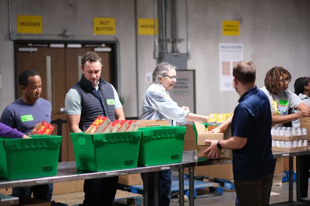 A group of people stand at a long table in a warehouse, packing food items such as cereal boxes and canned goods into green bins and cardboard boxes. Signs on the wall label sections for veggies, nut butter, and water.