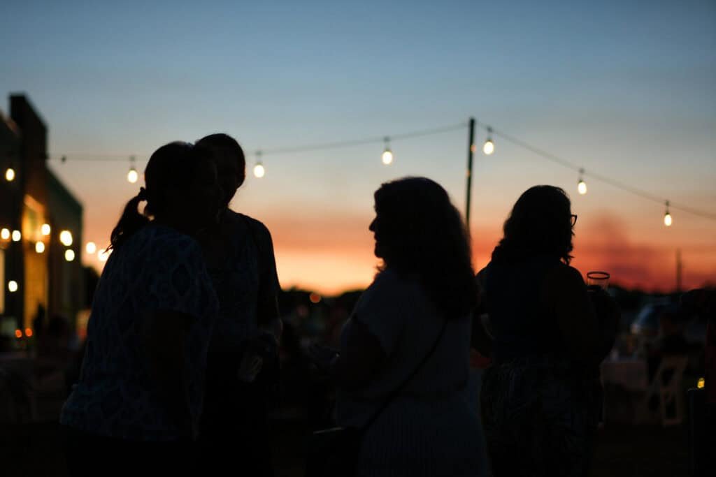 Silhouettes of four people talking outdoors at dusk, with string lights glowing above them and a colorful sunset sky in the background.