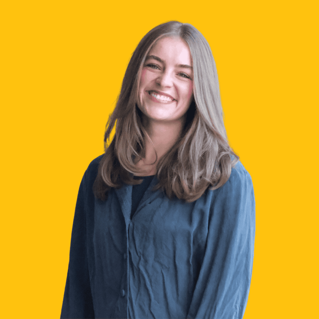 A young woman with long brown hair smiles warmly, wearing a blue button-up shirt against a bright yellow background.