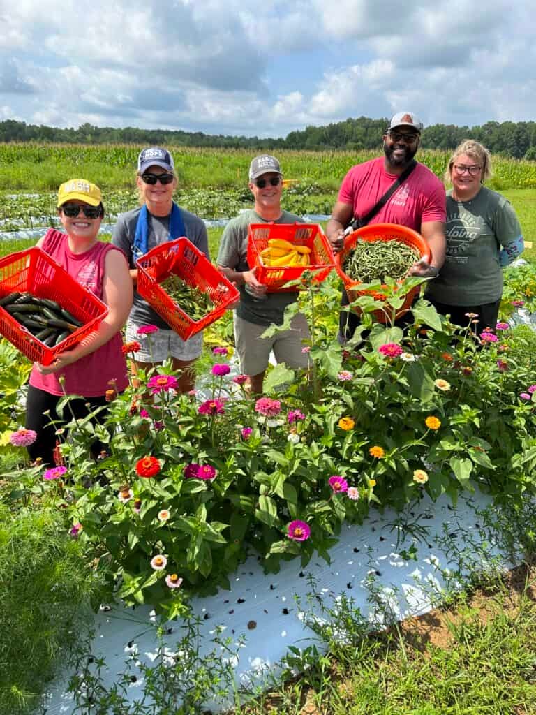 Five people stand in a flower-filled garden, smiling and holding red baskets of freshly harvested vegetables under a partly cloudy sky. Fields and trees are visible in the background.