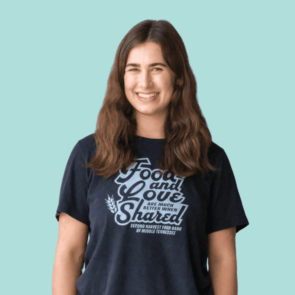 A young woman with long brown hair smiles at the camera. She is wearing a navy blue t-shirt that reads, Food and love are much better when shared. Second Harvest Food Bank of Middle Tennessee.