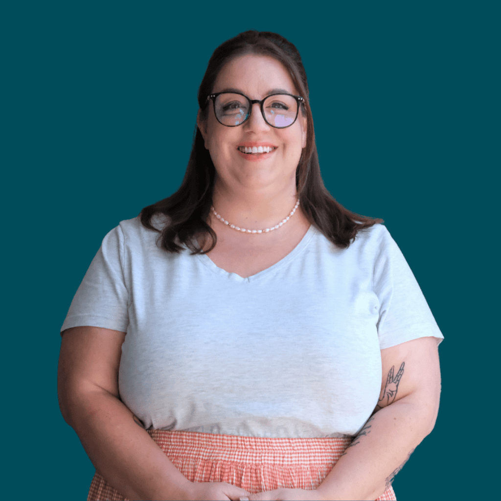 A woman with long brown hair, glasses, and a pearl necklace smiles at the camera. She is wearing a light gray top and a pink plaid skirt, standing in front of a solid teal background. A tattoo is visible on her left forearm.