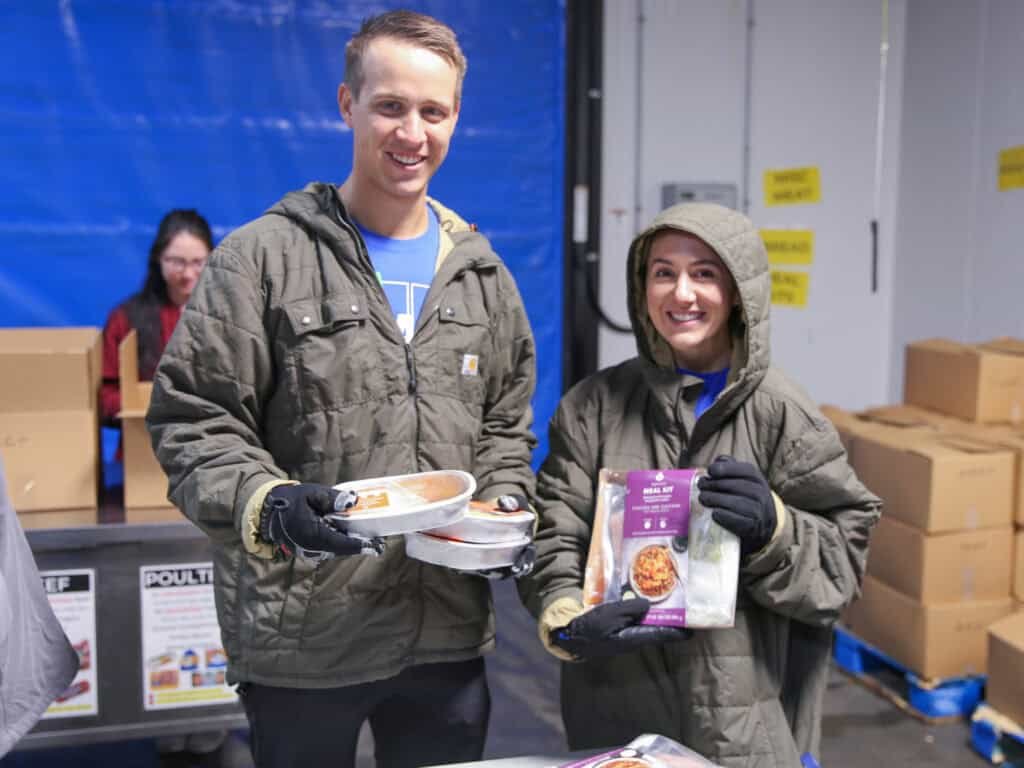 Two people wearing jackets and gloves smile while holding packaged food items at a food distribution center. Boxes and another volunteer working at a table are visible in the background.