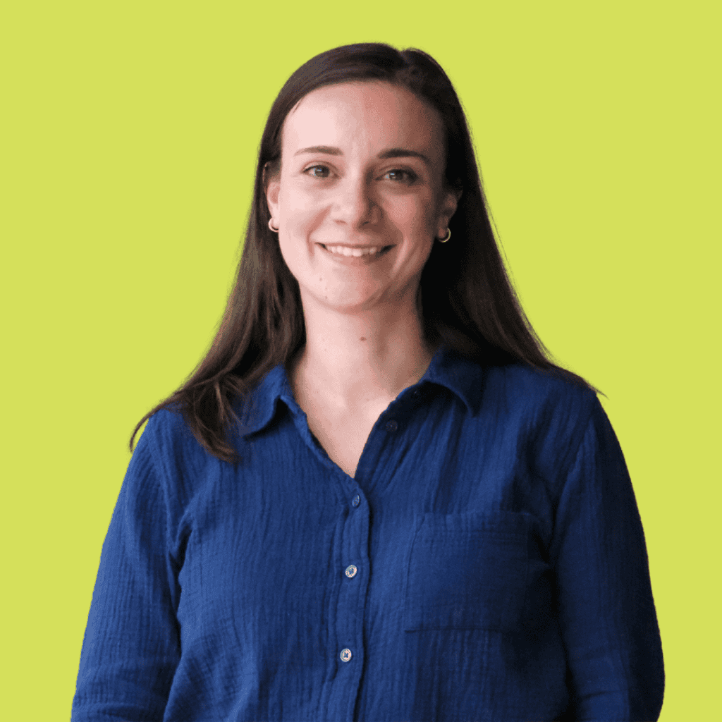A woman with long brown hair, wearing a blue button-up shirt, smiles at the camera while standing in front of a plain light green background.