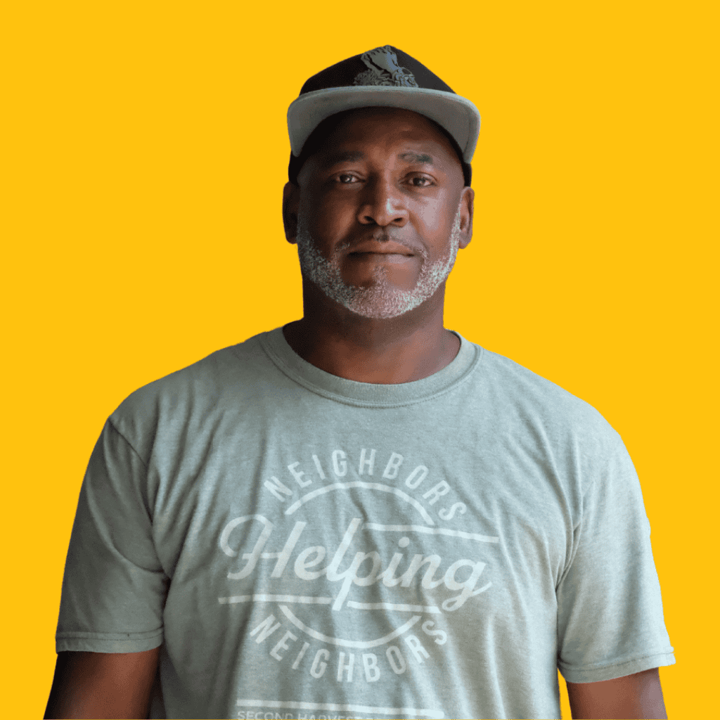 A man wearing a gray Helping Neighbors t-shirt and a baseball cap stands in front of a solid yellow background, looking at the camera with a neutral expression.