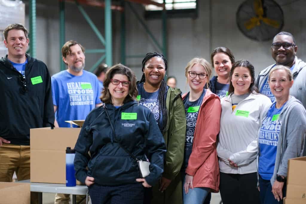 A diverse group of nine smiling adults poses together inside a warehouse, some wearing jackets or T-shirts, standing in front of cardboard boxes and metal shelves, suggesting a volunteer or community event.