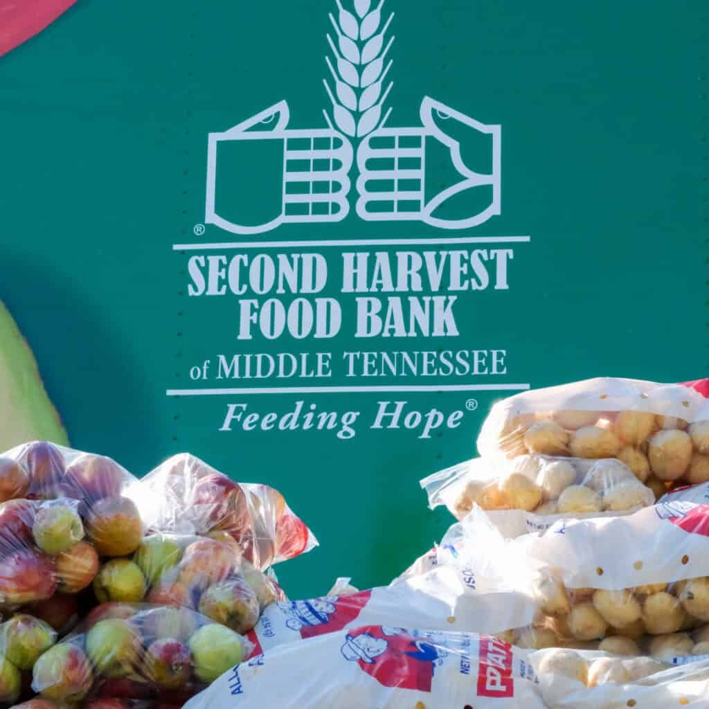 Bags of apples and potatoes are stacked in front of a green sign for Second Harvest Food Bank of Middle Tennessee, featuring a logo with two hands holding wheat and the slogan Feeding Hope.