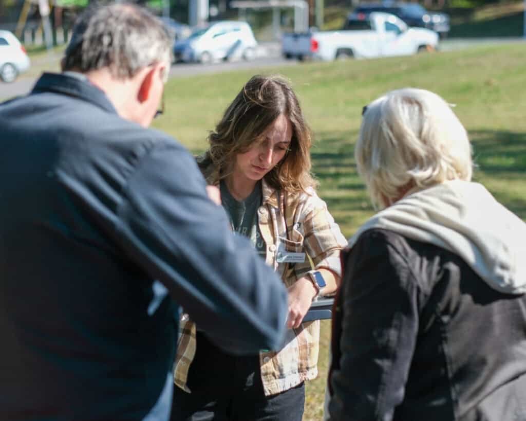 A woman outdoors looks at her smartwatch while two older adults stand nearby, partially turned away from the camera. Parked cars and a grassy area are visible in the background.