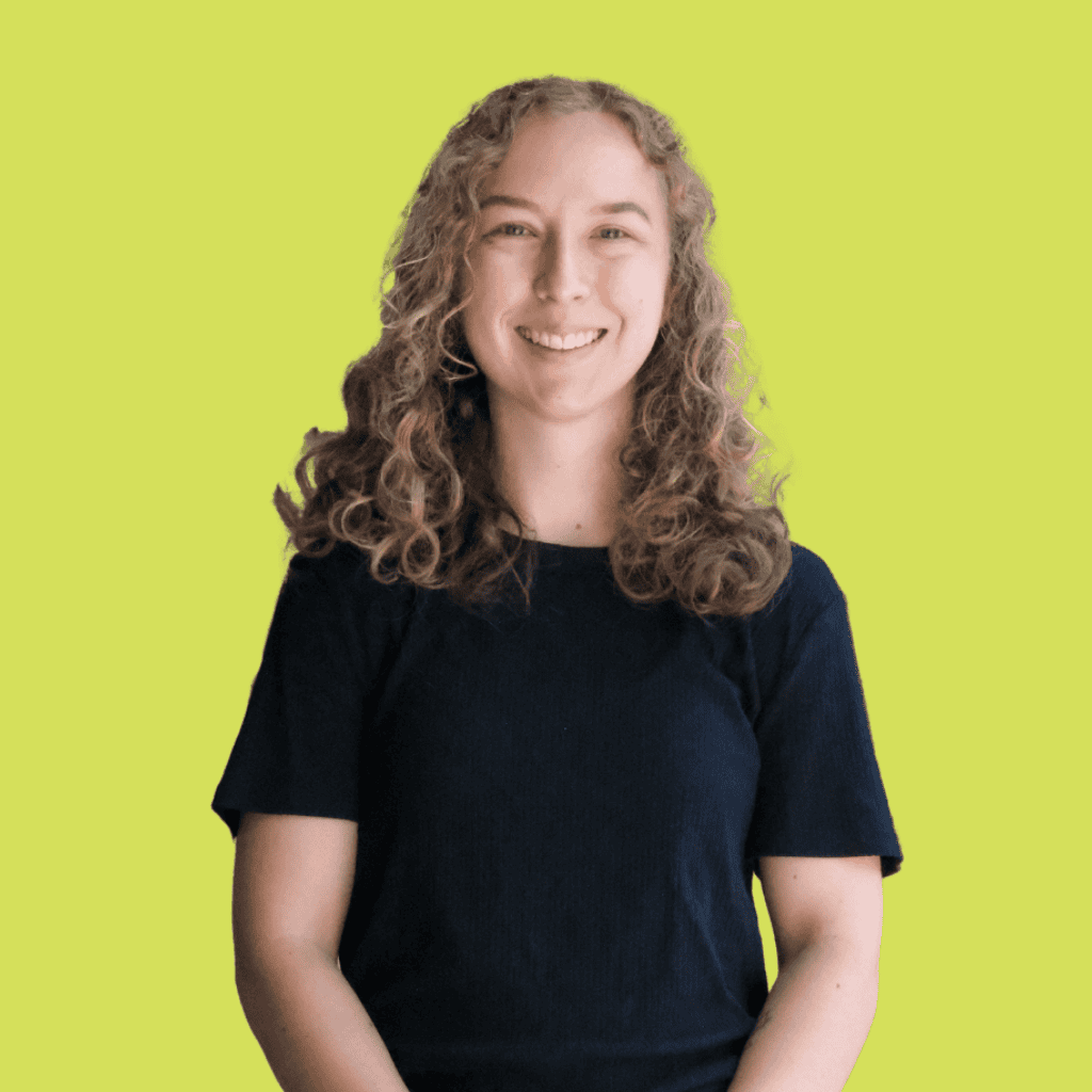 A person with curly light brown hair, wearing a short-sleeved black shirt, smiles while standing against a solid light green background.
