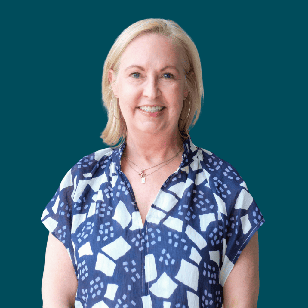 A smiling woman with short blond hair wearing a blue and white patterned blouse stands against a solid teal background.