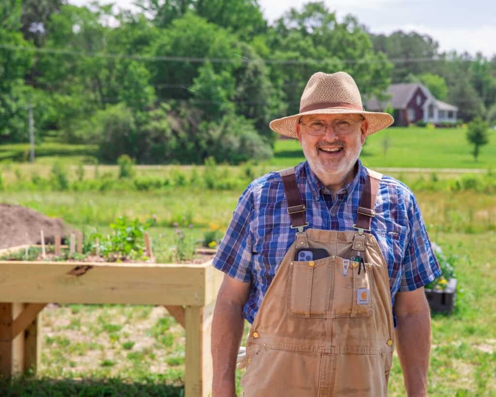 A smiling older man in overalls and a straw hat stands outdoors near a raised garden bed, with greenery and a red house in the background.