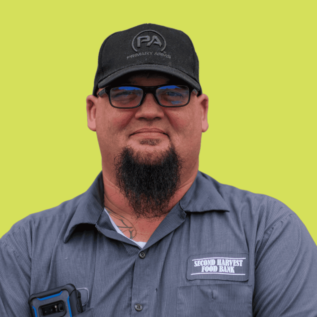 A man with a long goatee, glasses, and a black cap stands against a yellow background. He wears a gray shirt with a Second Harvest Food Bank patch and holds a phone in his pocket.