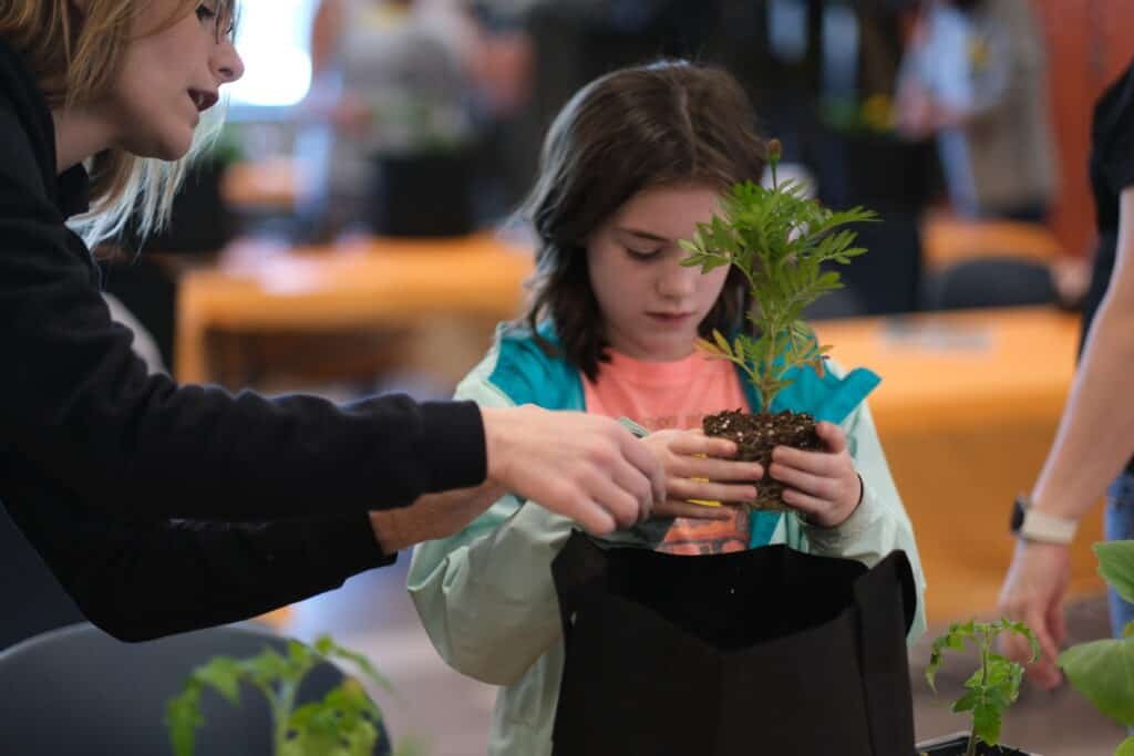 A young girl holds a small plant while an adult helps her prepare to plant it in a black fabric pot. Tables with orange tablecloths and other plants are visible in the background.