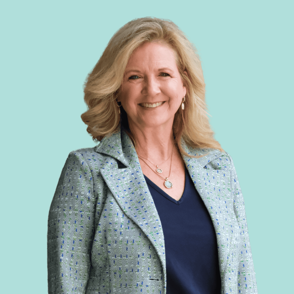 A woman with blonde hair smiles, wearing a patterned light blue blazer over a navy top, with layered necklaces, standing against a solid light blue background.