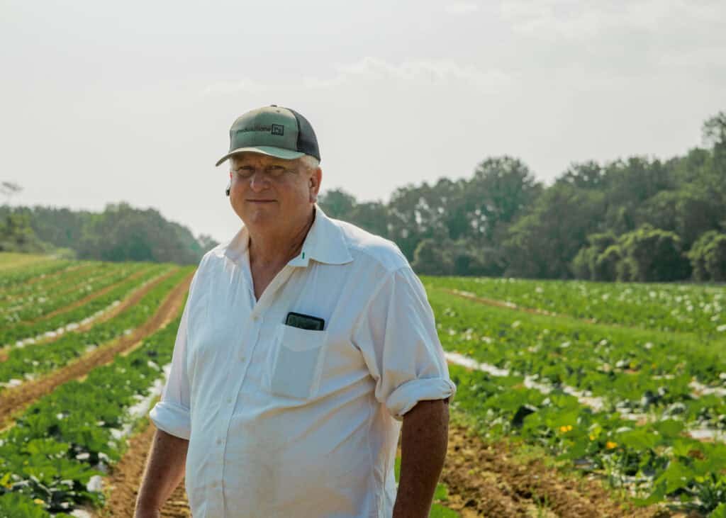 A man in a white shirt and cap stands in a green crop field with rows of plants stretching into the distance, trees and a bright sky in the background.