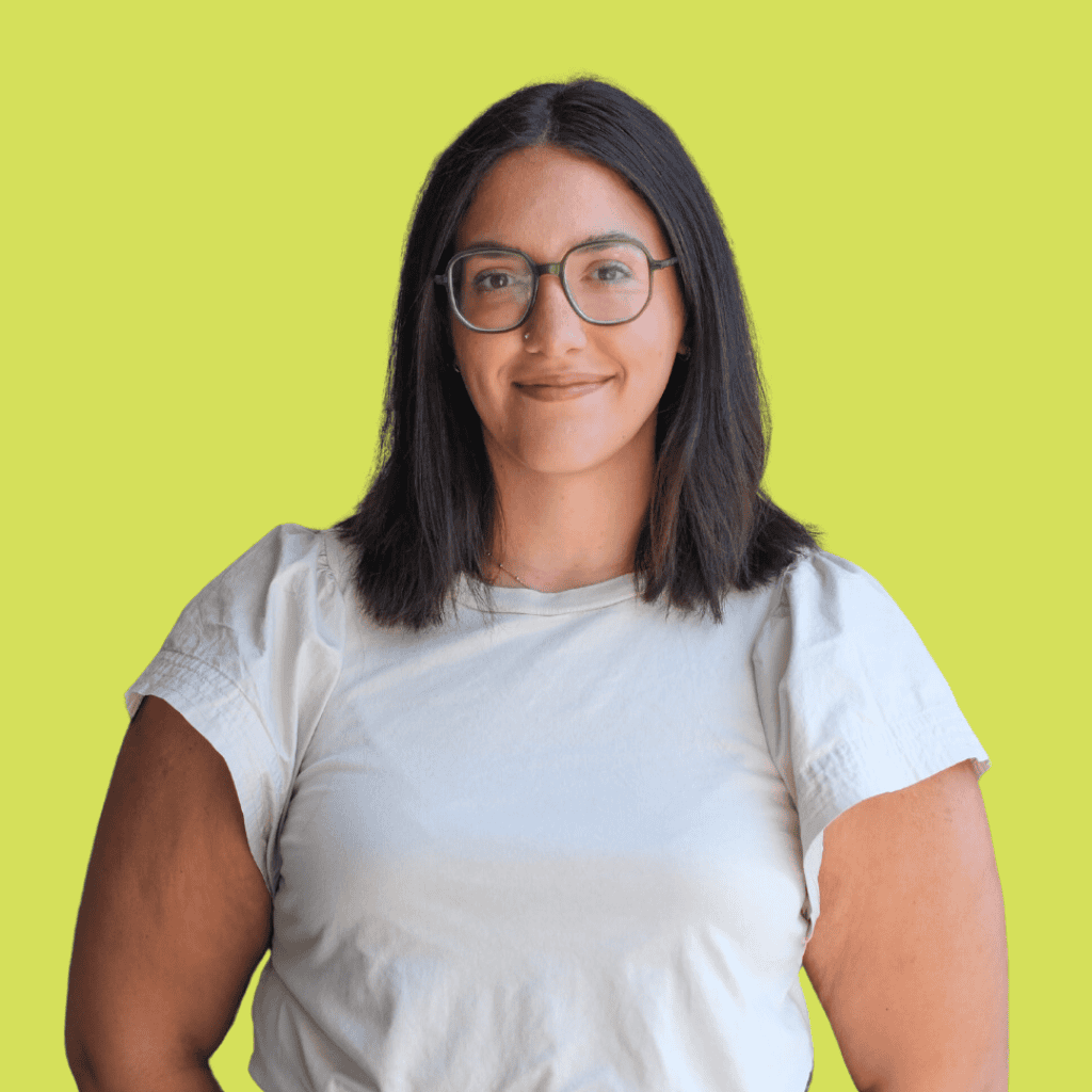 A woman with straight dark hair and glasses, wearing a white short-sleeve shirt, stands smiling in front of a solid light green background.