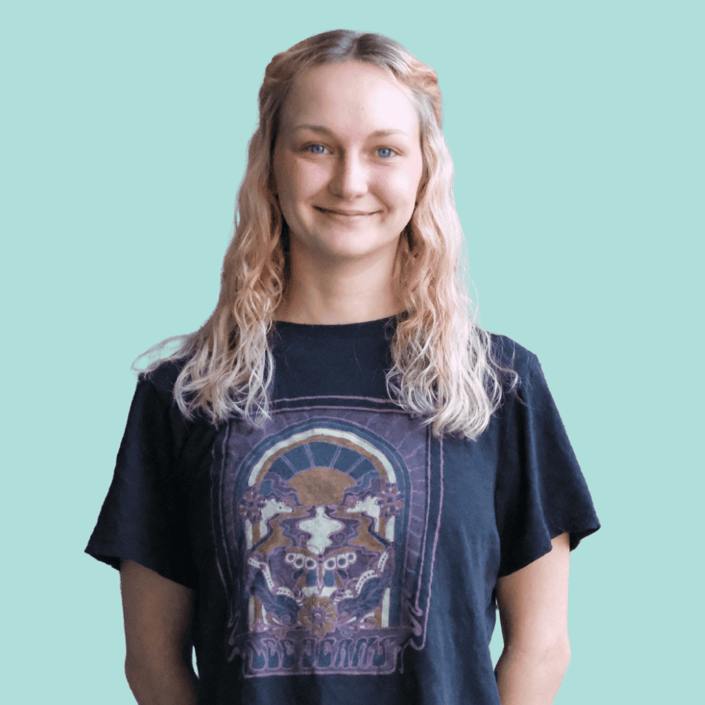 A person with wavy, light blonde hair is standing against a pale teal background, smiling. They are wearing a black graphic t-shirt featuring a colorful, intricate design.
