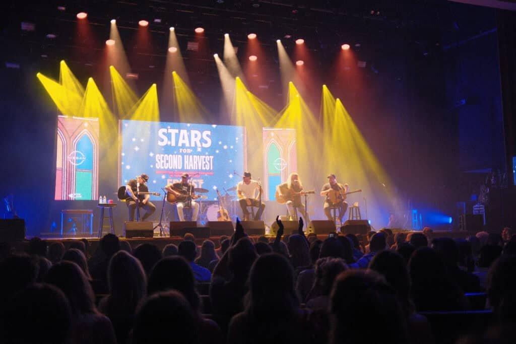 A band of six musicians performs onstage with acoustic guitars under yellow stage lights, in front of a screen that reads Stars for Second Harvest. Audience members are visible in the foreground.