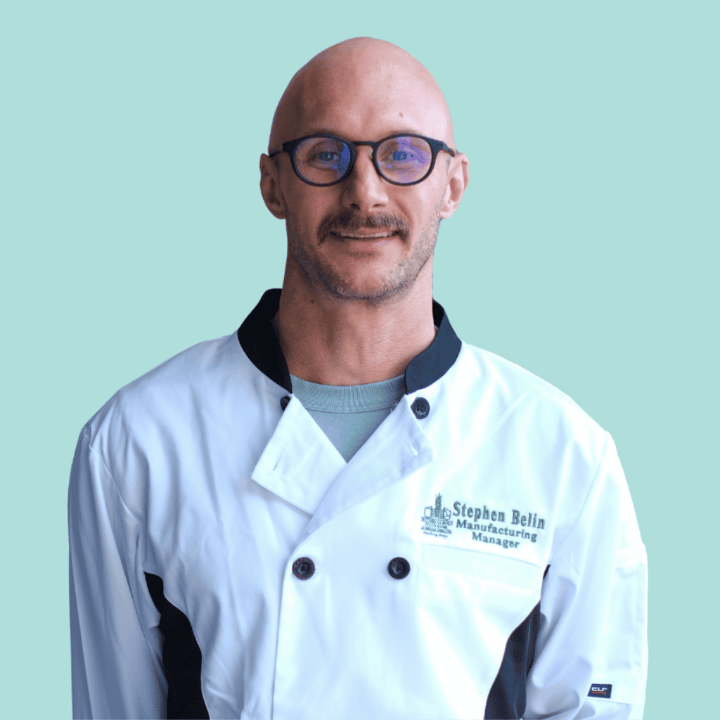 A man with a shaved head, glasses, and a short beard wears a white chef jacket with black trim. The text on the jacket reads, Stephen Belin, Manufacturing Manager. The background is a solid light blue.