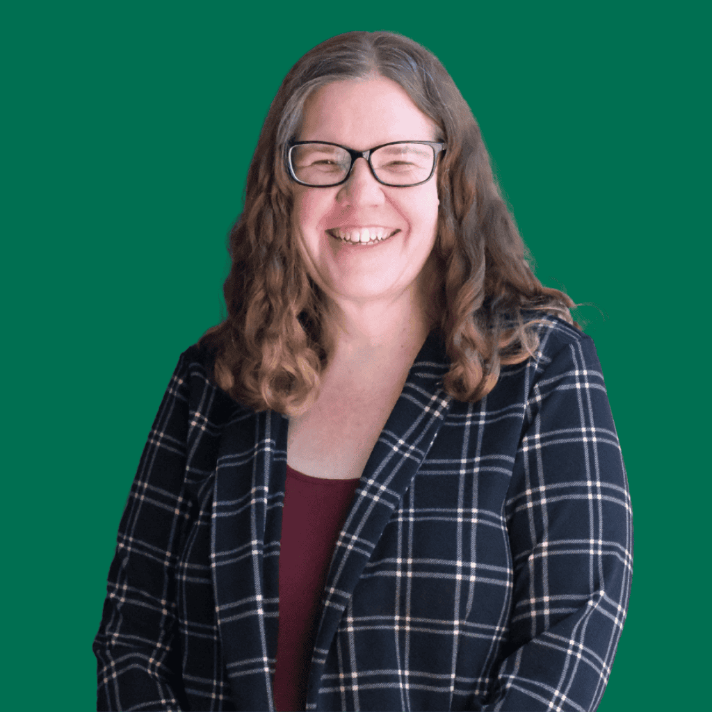 A woman with long, wavy brown hair and glasses is smiling. She is wearing a dark plaid blazer over a maroon top, standing against a solid green background.