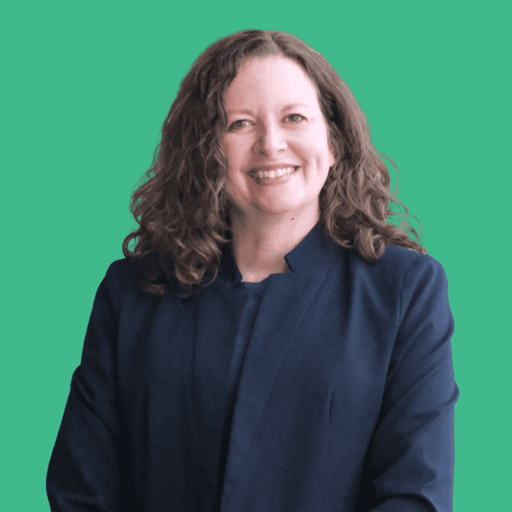 A woman with curly brown hair wearing a navy blazer smiles at the camera against a solid green background.